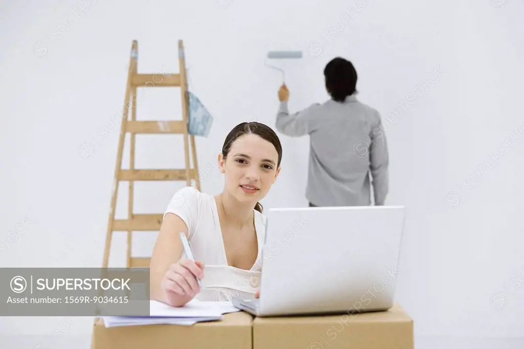 Young woman sitting at makeshift desk, using laptop computer, smiling at camera, man painting wall in background