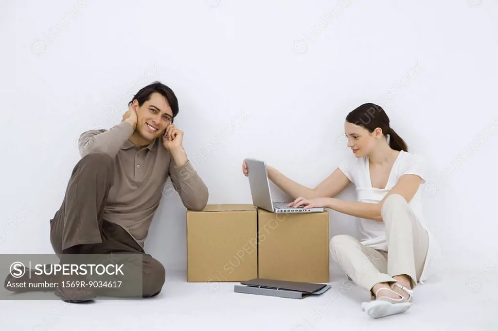 Couple sitting at makeshift desk, woman using laptop computer, man using cell phone and smiling at camera