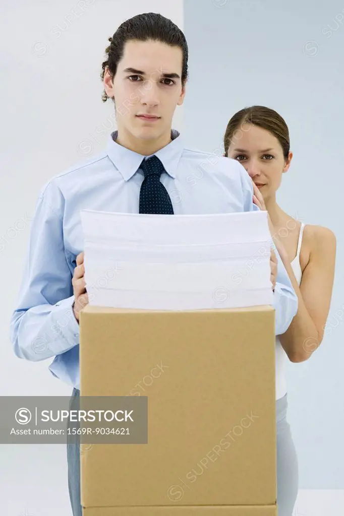 Young professional with documents stacked on cardboard box, woman standing behind him with her hand on his shoulder, both looking at camera