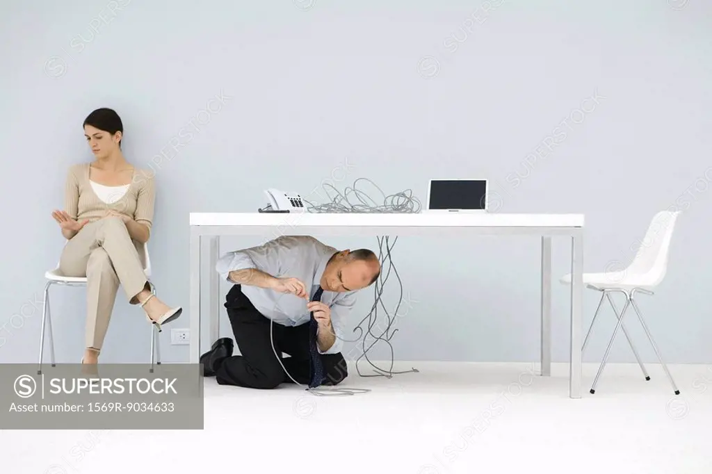 Man kneeling under desk, connecting tangled wires, woman sitting in chair, looking at hand