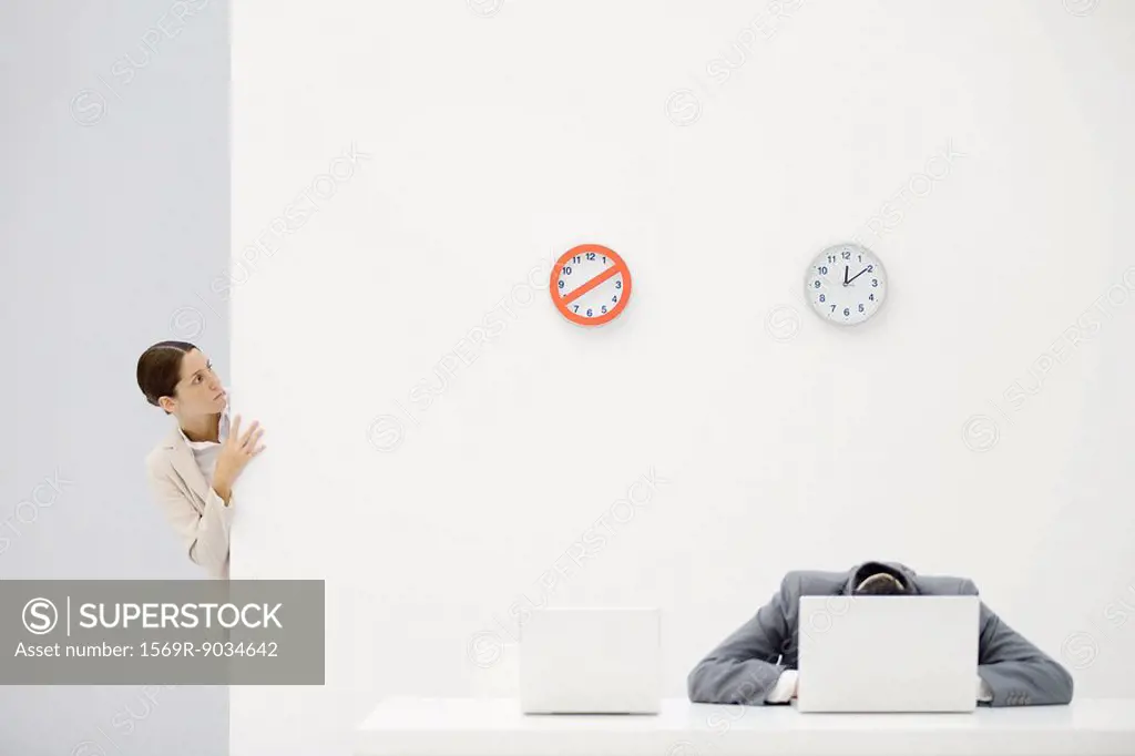 Female office worker peeking around corner at clocks on wall, colleague sitting in front of laptop with head down