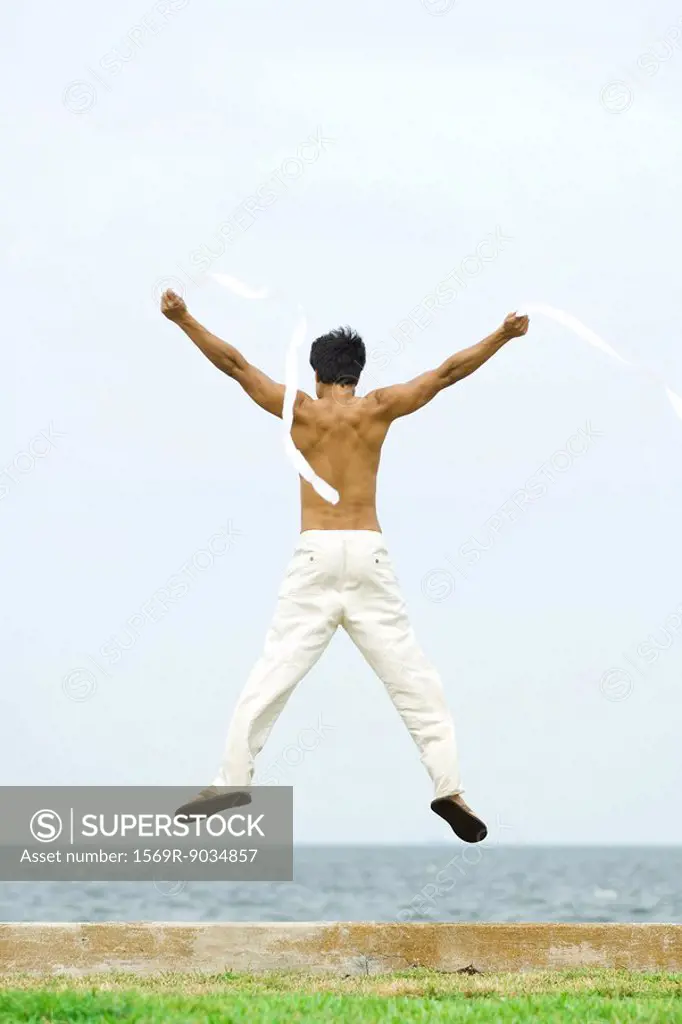 Man jumping into the air with streamers in hands, rear view, ocean horizon in background