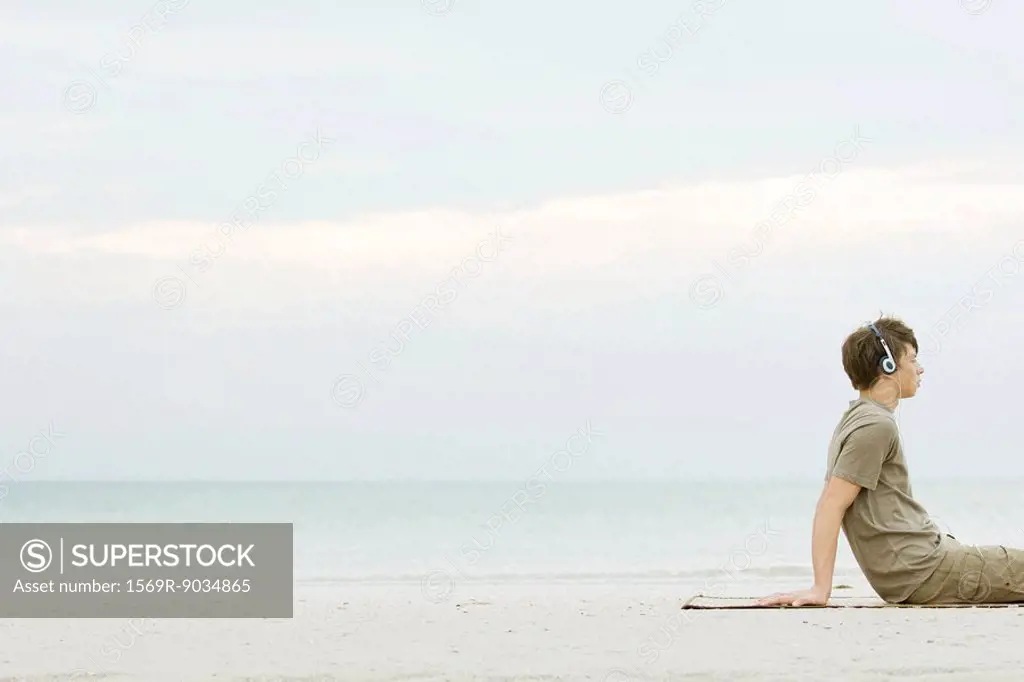 Teen boy sitting on beach listening to headphones, cropped
