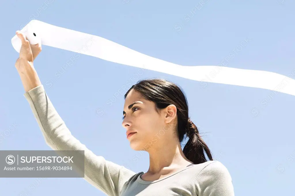 Woman holding up toilet paper roll outdoors, paper streaming in wind, low angle view