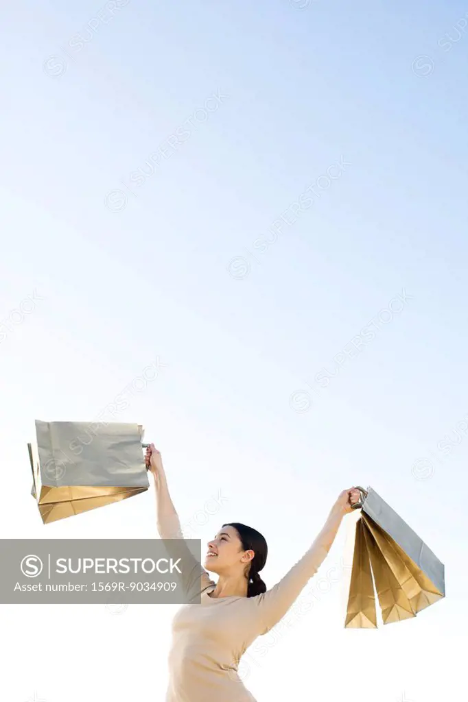 Woman holding up shopping bags, smiling, low angle view