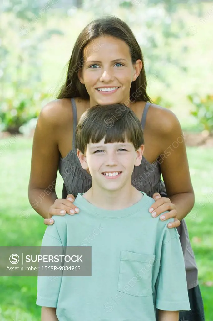 Teen girl standing behind younger brother, resting her chin on his head, both smiling at camera