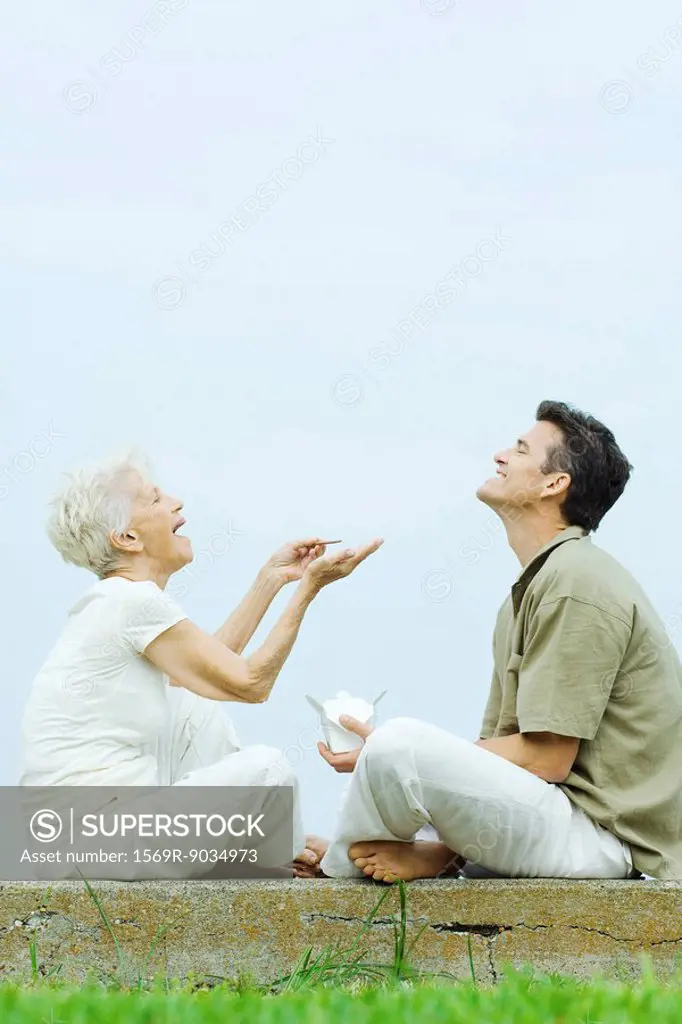 Senior woman and adult son sitting face to face on the ground, sharing takeout food, laughing