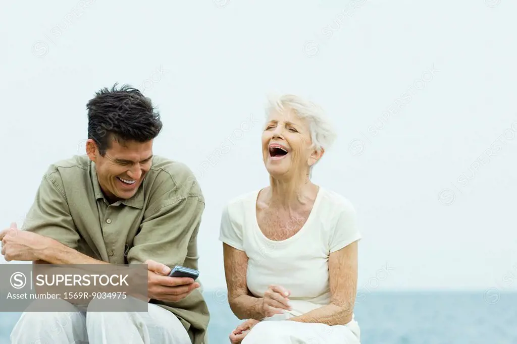 Senior woman and adult son sitting side by side outdoors, laughing, man holding cell phone