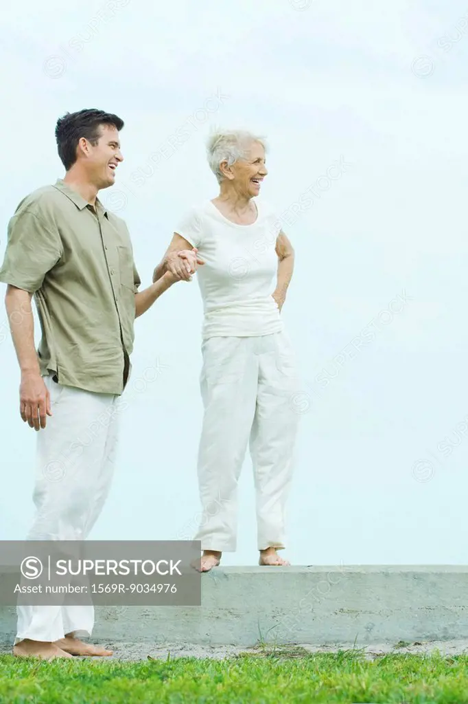 Senior woman standing on ledge, holding adult son´s hand, both looking away, smiling