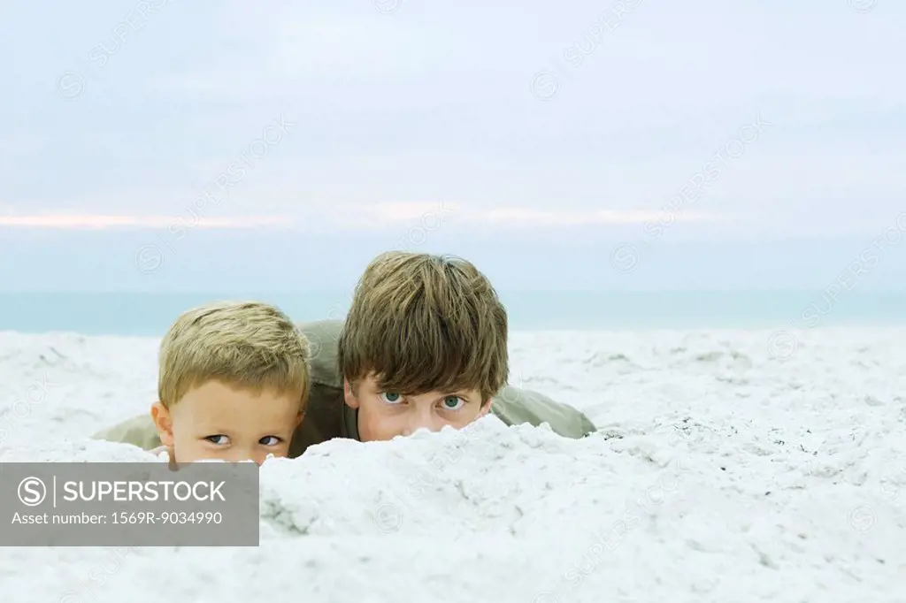 Teen boy and little brother at the beach, peeking over sand at camera