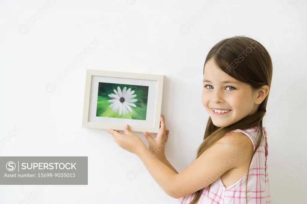 Little girl holding framed picture against wall, smiling over shoulder at camera