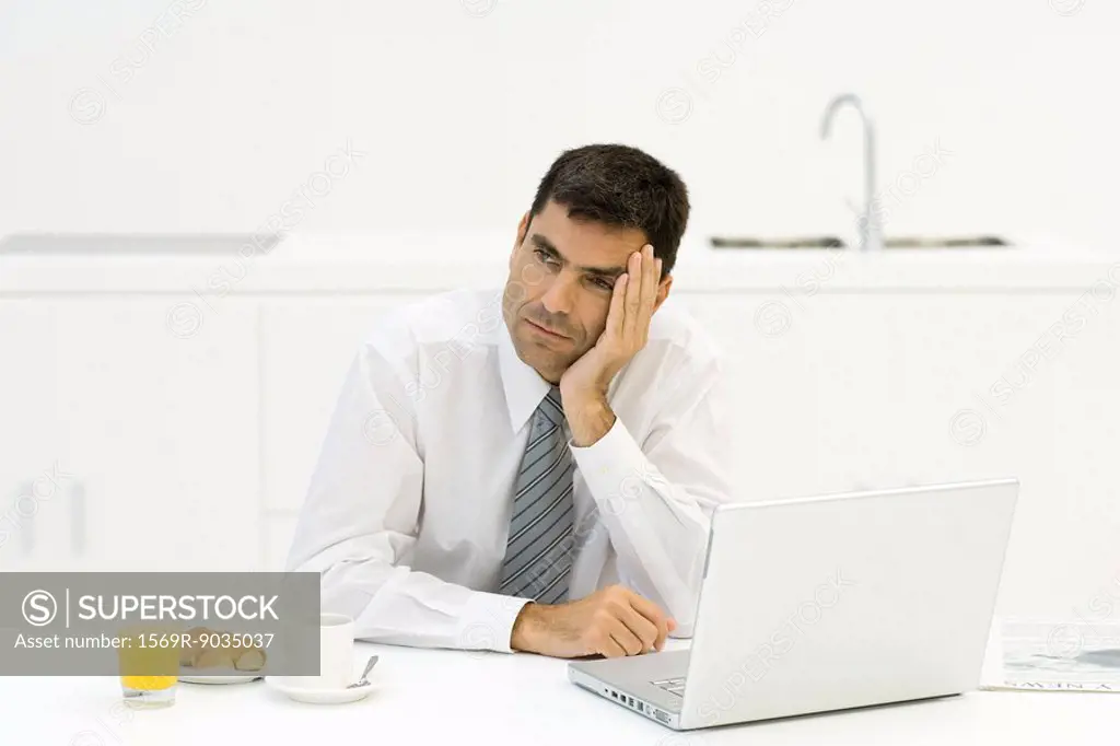 Businessman sitting in kitchen with breakfast and laptop, leaning head on hand, looking away