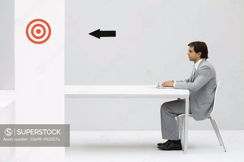 Man sitting at desk, using laptop computer, arrow on wall pointing at bullseye