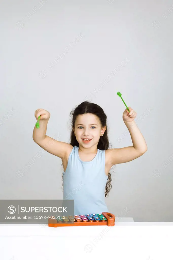 Little girl playing xylophone, arms raised, looking at camera