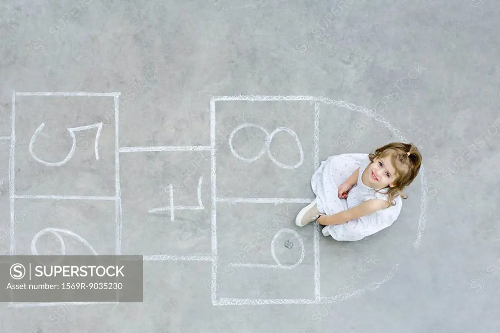 Girl sitting down on hopscotch grid, looking up, overhead view