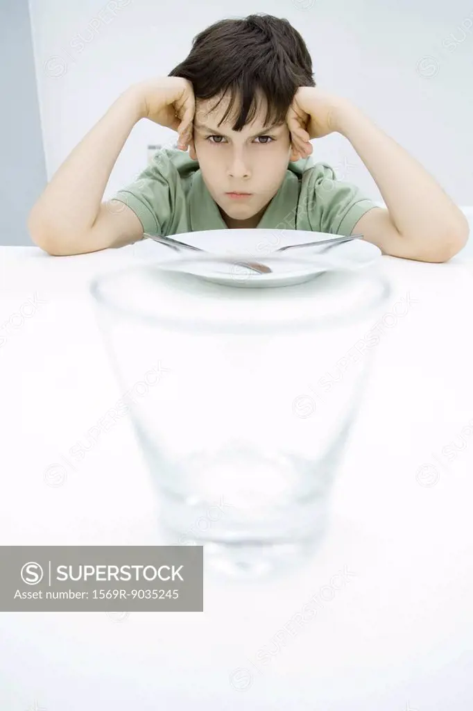 Boy sulking at kitchen table, leaning on elbows, empty glass in foreground