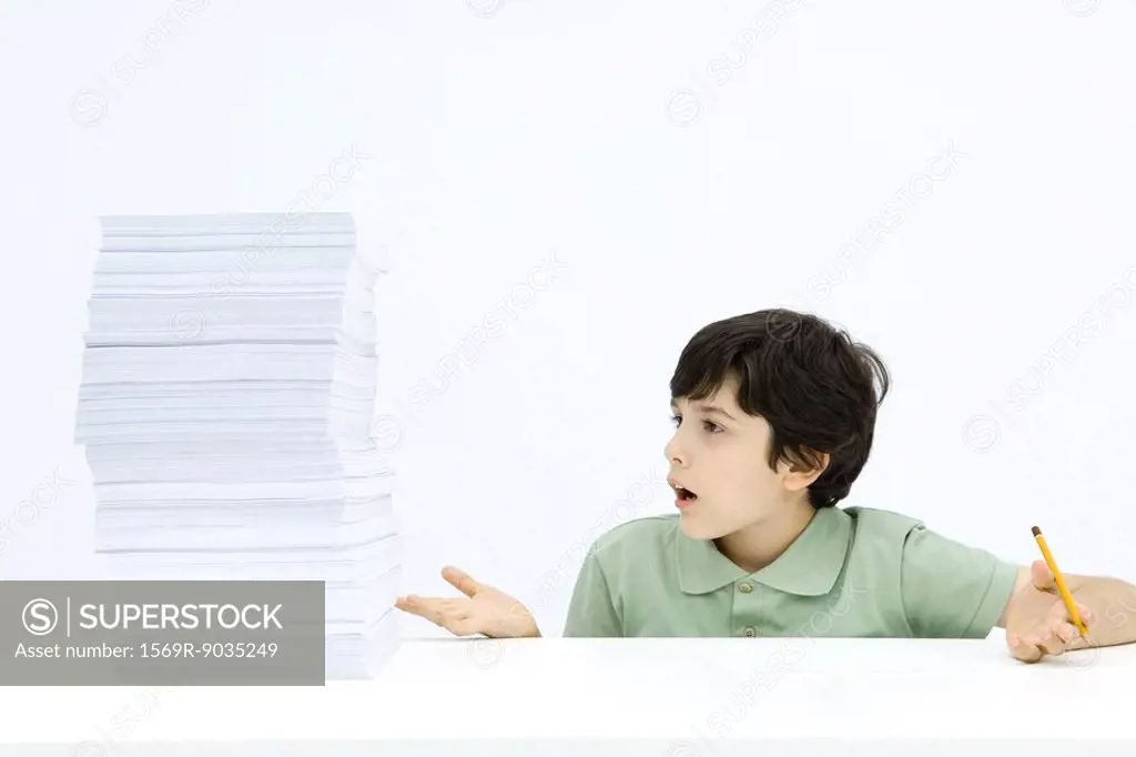 Boy looking at tall stack of paper, shrugging in disbelief, holding pencil