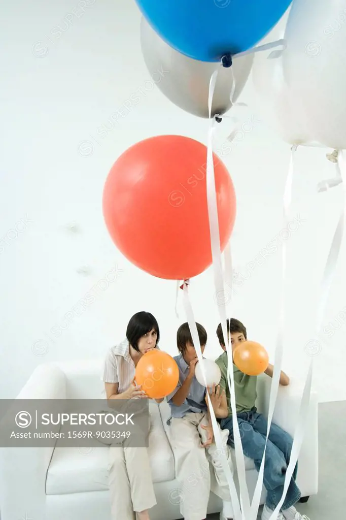 Mother and two sons sitting on sofa, inflating balloons
