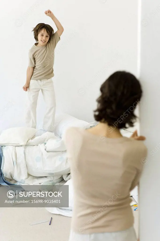 Little boy jumping on unmade bed, mother watching from doorway
