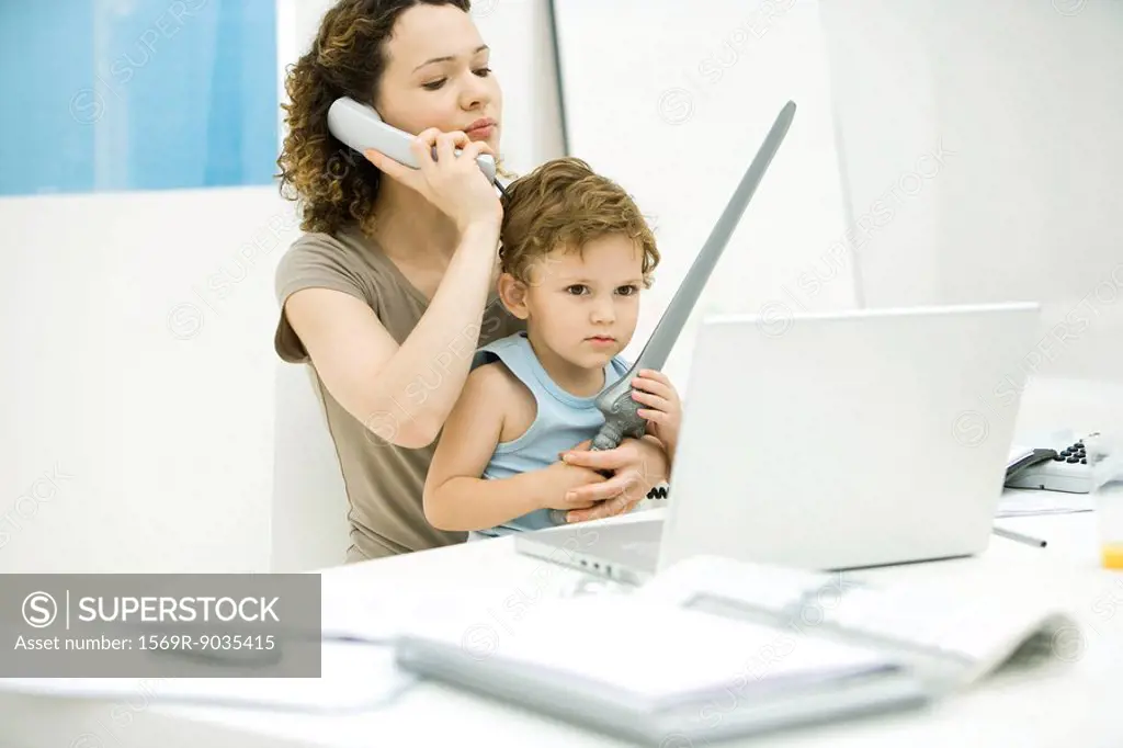 Young mother sitting at desk using phone while son sits on lap, holding sword