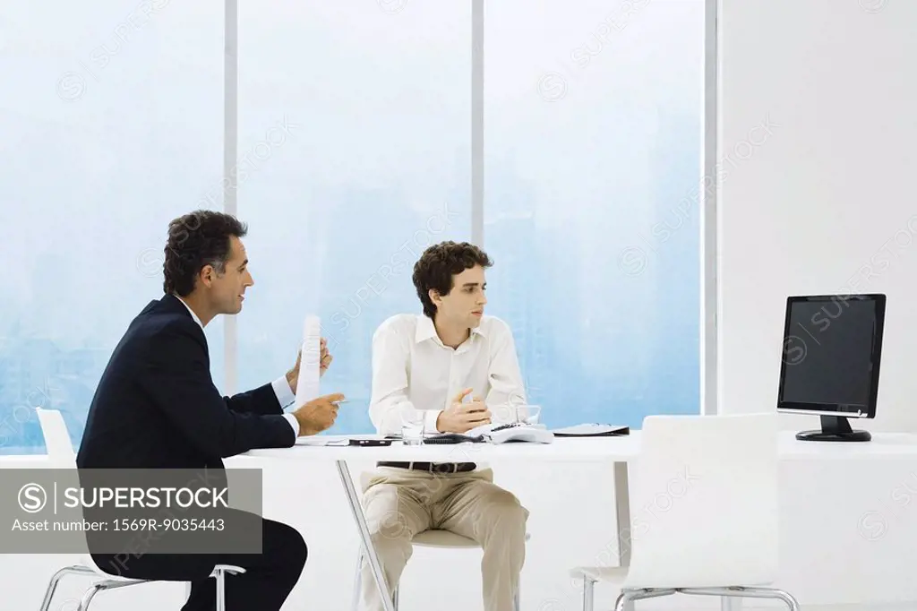 Businessmen sitting at table, looking at monitor, teleconferencing