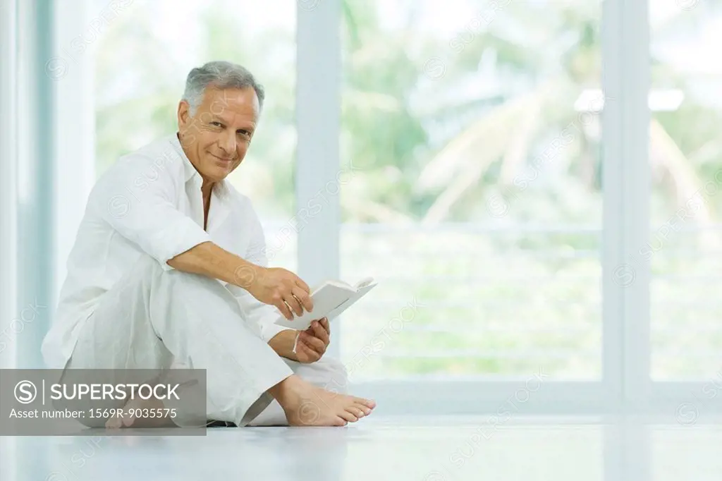 Mature man sitting on the floor, holding book, smiling at camera