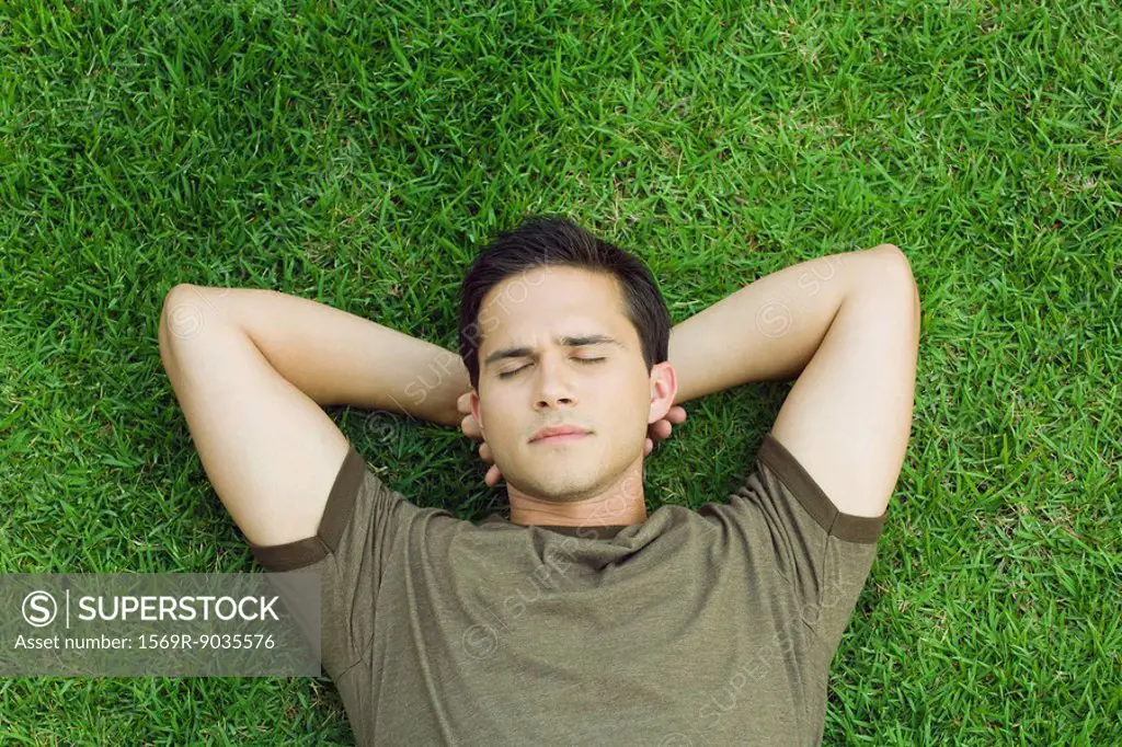 Young man lying on grass with hands behind head, eyes closed, high angle view