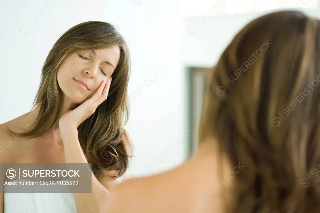 Woman touching face, eyes closed, reflected in mirror