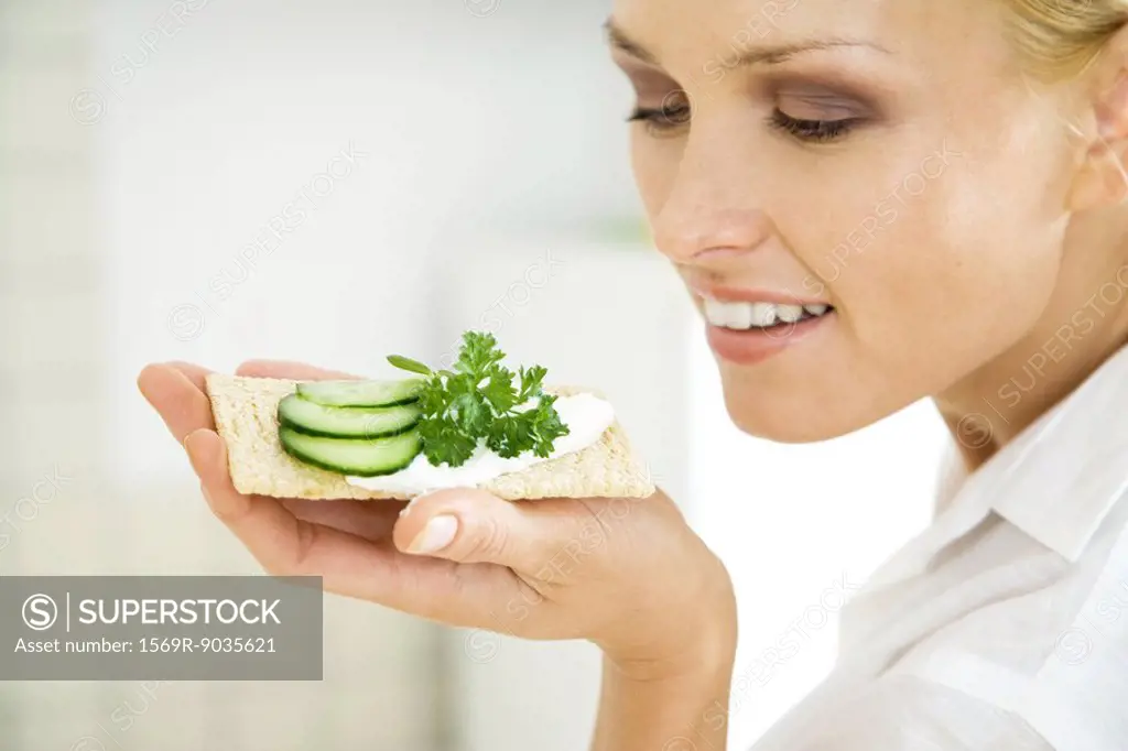 Woman holding up cracker with cucumber slices, smiling