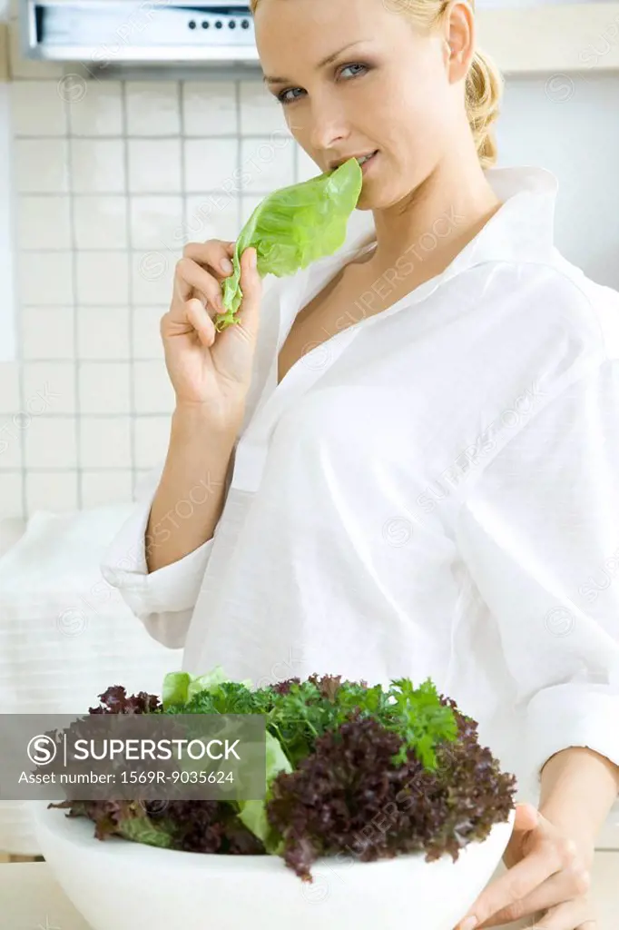 Woman standing in front of large salad bowl, eating lettuce, looking at camera