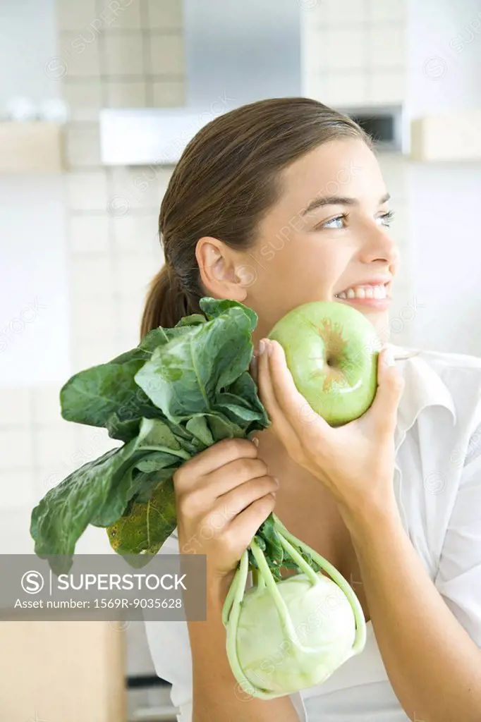Woman holding apple and kohlrabi, looking away, smiling