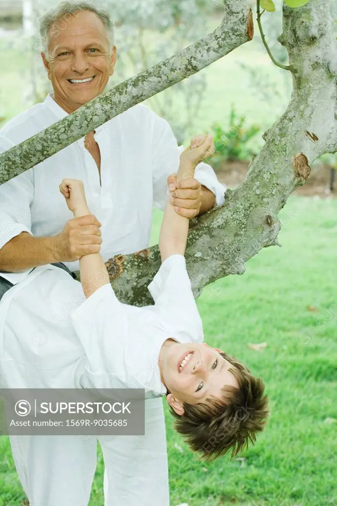 Grandfather holding up grandson hanging from tree, both smiling at camera