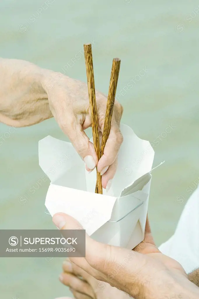 Male hand holding container, senior woman´s hand reaching in with chopsticks, cropped