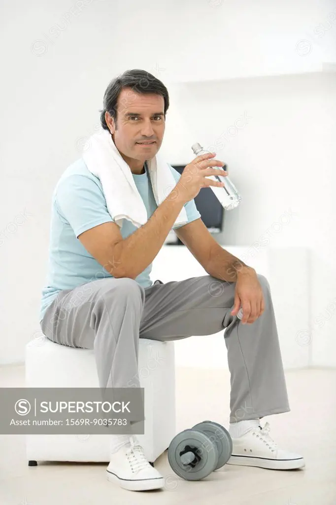 Man in sports clothing holding water bottle, dumbbell at his feet