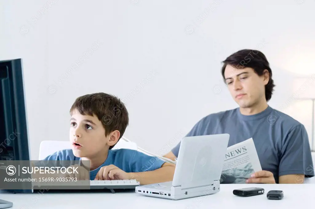 Little boy computer and portable DVD player, father reading newspaper in background