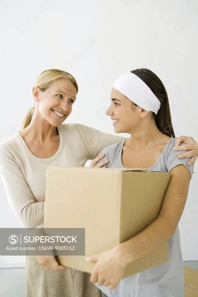Mother with arm around daughter´s shoulder, daughter holding cardboard box, both smiling
