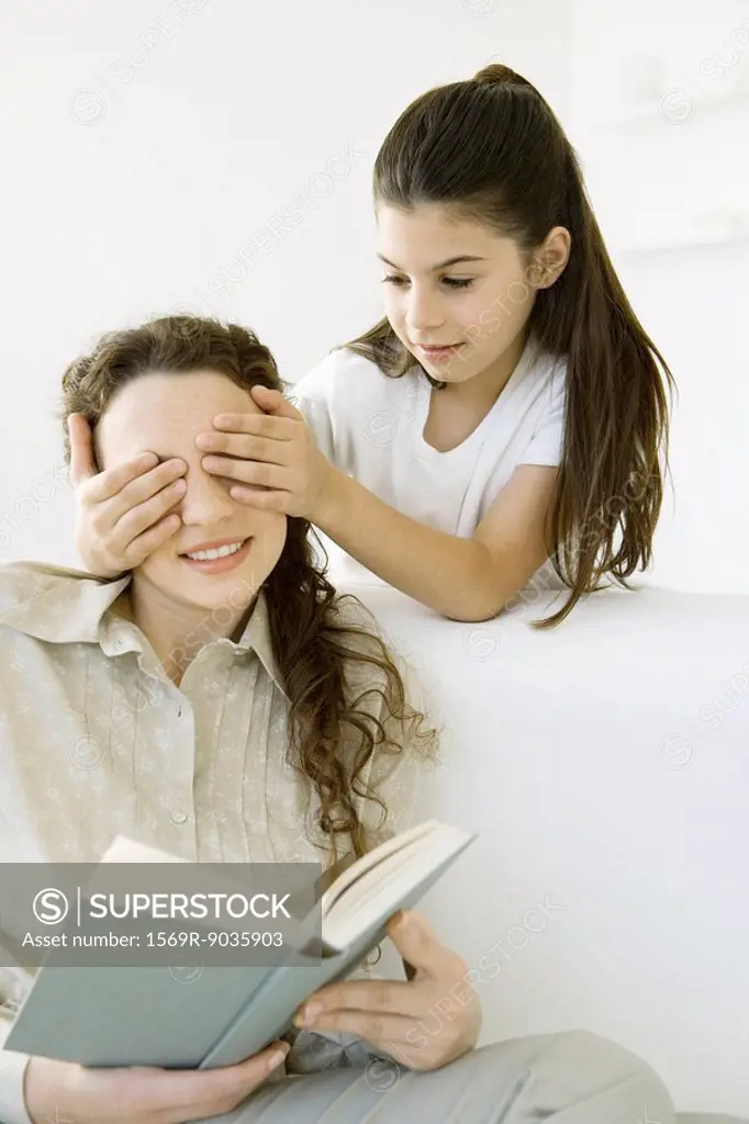 Girl standing with hands over her mother´s eyes, woman holding book