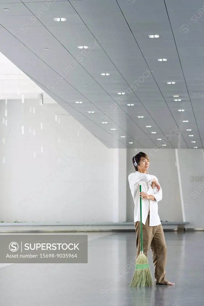 Man standing in lobby, resting arm on broom, listening to headphones