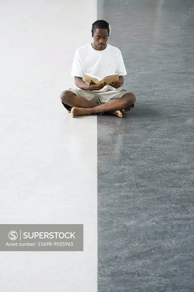 Teenage boy sitting cross-legged on the floor, reading a book, listening to headphones