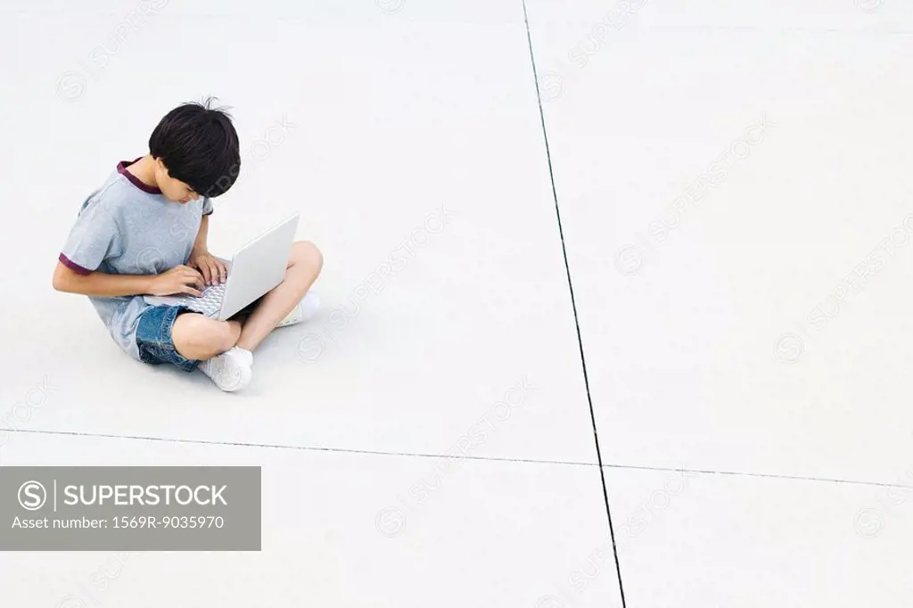 Boy sitting cross-legged on the ground, using laptop, high angle view