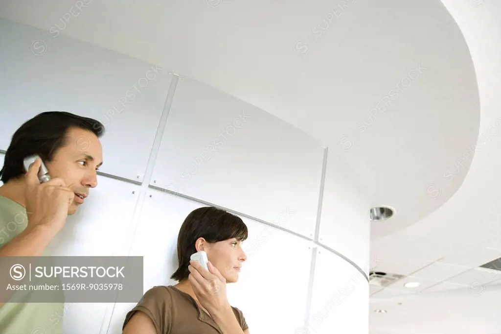 Man and woman indoors, looking away, both holding cell phone to ear, cropped