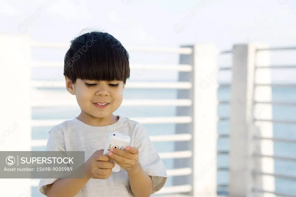 Little boy outdoors, looking down at cell phone, smiling