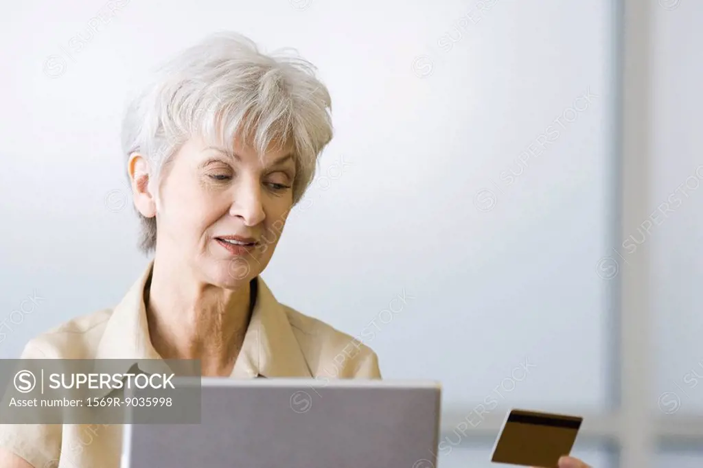 Senior woman looking at credit card, sitting behind laptop