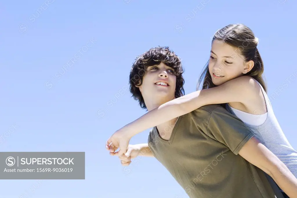 Teen boy carrying younger sister on his back, both smiling at each other, low angle view