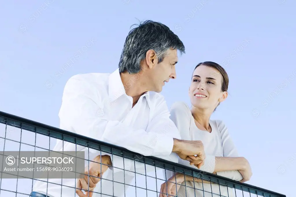 Couple leaning on wire fence, smiling at each other, low angle view