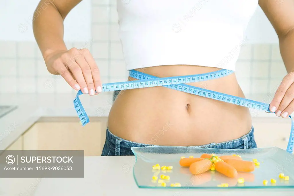 Woman wrapping a measuring tape around her stomach, plate of vegetables on table