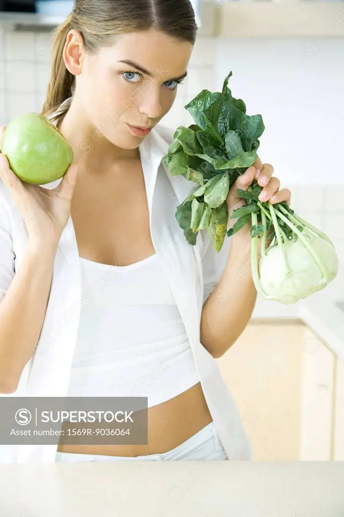 Woman holding up fresh kohlrabi and apple, looking at camera