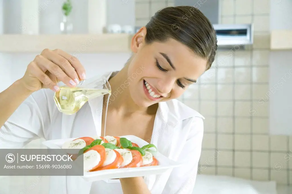 Young woman pouring olive oil over tomato mozzarella salad, smiling