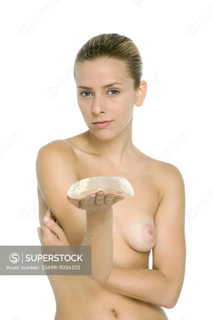 Nude woman holding breast implant in hand, looking at camera