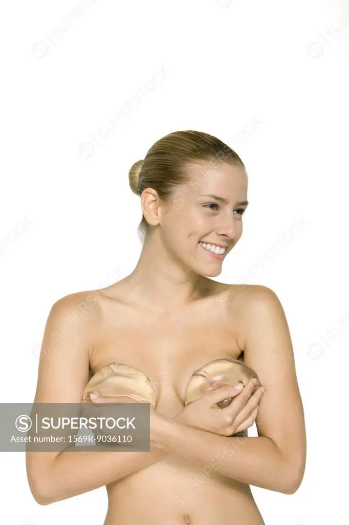 Nude woman holding breast implants over her chest, smiling